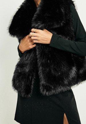 Donna che indossa un gilet senza maniche in pelliccia sintetica nera sopra una maglia a maniche lunghe, mentre tiene il colletto del gilet con le dita adornate da anelli d'oro.