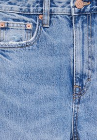 Jean en denim bleu clair en gros plan montrant la poche avant, les passants de ceinture, les rivets en cuivre et le bouton métal avec des détails de couture.
