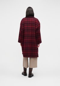Red and black plaid wool coat with a relaxed fit, featuring a collar and long sleeves, paired with beige trousers and brown boots.