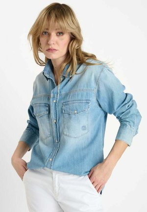 Chemise en denim bleu clair à manches longues, bouton devant, deux poches poitrine et une finition légèrement délavée. Coupe structurée avec un matériel minimal.