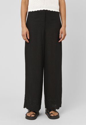 Femme portant un pantalon noir à jambes larges avec ourlet ajouré floral, top blanc sans manches en maille, sandales noires et bagues dorées aux doigts.