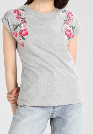 Woman wearing a light gray T-shirt with pink and red floral embroidery on the shoulders and light blue denim jeans.