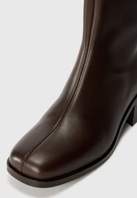 Bottine en cuir brun avec un bout carré et des détails de couture subtils. Le talon est épais et la semelle est plate pour plus de stabilité.