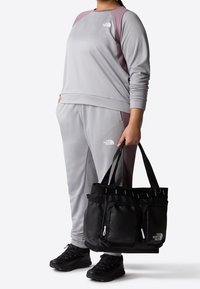 Completo sportivo grigio con accenti viola e una finitura tessurizzata, abbinato a una borsa a tracolla nera con più tasche e un logo.