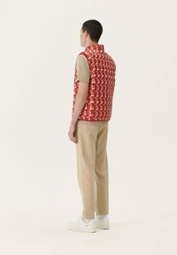 Gilet imbottito rosso con un motivo bianco ripetuto, indossato sopra una camicia beige, abbinato a pantaloni leggeri beige e sneaker bianche.