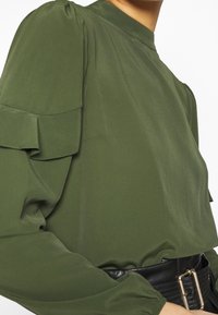 Mörkgrön blus med volangärmar, hög krage och en slät yta, i kombination med ett svart bälte med en rektangulär spänne.