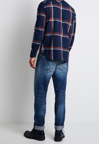 Man som bär en mörkblå, röd och vit rutiga skjorta, urblekta blå jeans med rullade fållar och svarta läderskor, står mot en vit vägg.