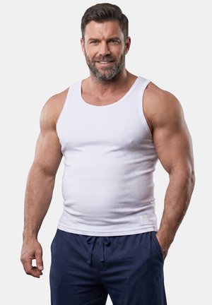 Muscular man smiling wearing a white sleeveless tank top and navy blue drawstring pants with one hand in pocket.