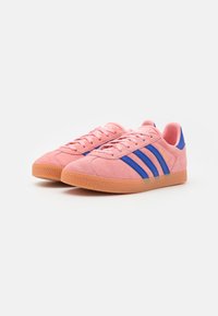 Pink suede sneakers with blue stripes, textured gum rubber sole, circular tread pattern, and lace-up front. Classic athletic design.