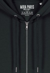 Hoodie di cotone verde scuro con zip YKK, cordini regolabili e un'etichetta con la scritta "MIRA PARIS" e istruzioni per la cura all'interno.