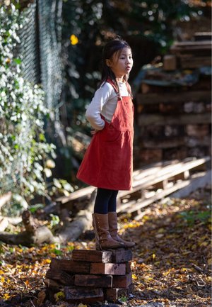 Rotes Corduroy-Latzkleid über einem cremefarbenen Langarmshirt, schwarzen Leggings und braunen kniehohen Stiefeln, steht auf Ziegelsteinen im Freien.