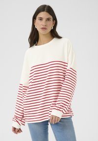 Dlhý rukáv v off-white farbe s horizontálnymi červenými pruhmi. Má rebrované manžety a voľný strih. V kombinácii s svetlomodrými džínsami.