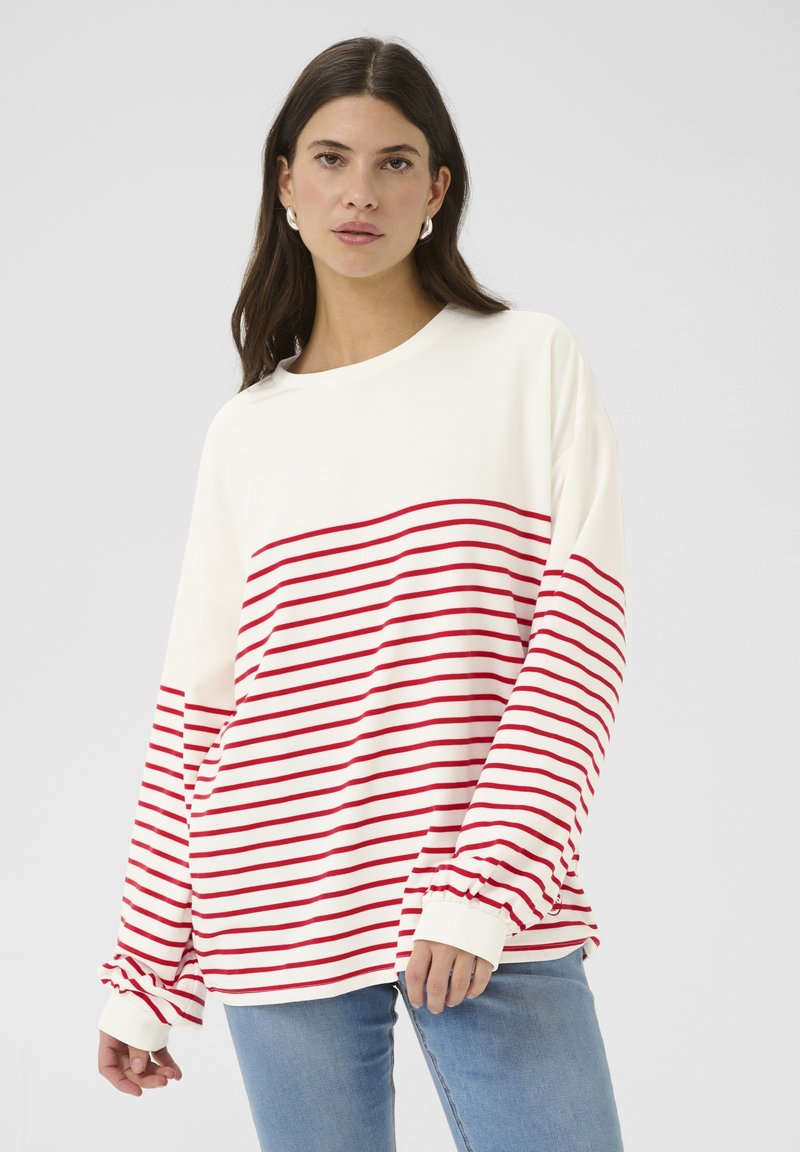 Dlhý rukáv v off-white farbe s horizontálnymi červenými pruhmi. Má rebrované manžety a voľný strih. V kombinácii s svetlomodrými džínsami.