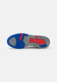 Semelle de chaussure avec une bande de roulement en caoutchouc gris, bleu et rouge présentant des zones d'adhérence texturées et des motifs géométriques sur fond blanc.