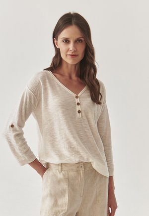 Maglione lungo beige chiaro a maniche lunghe con scollatura a V e tre bottoni decorativi sul davanti. Tessuto morbido e strutturato con maniche arrotolate e vestibilità rilassata.