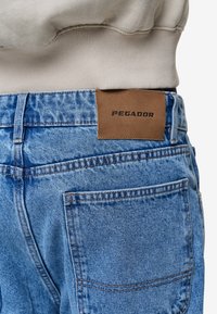 Svetle modré džíny z denimu s hnědou koženou nášivkou s označením "PEGADOR." Mají středně vysoký pas, standardní kapsy a viditelné detaily šití.