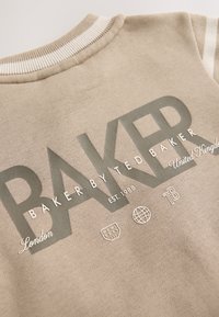 Sweat-shirt beige avec encolure côtelée et poignets rayés. Présente un grand graphisme "BAKER" en gris, ainsi que divers petits logos et textes.
