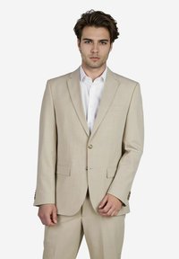 Beige maßgeschneiderter Anzug mit einreißiger Jacke und Revers. Gerade geschnittene Hose ergänzt die Jacke. Darunter ein weißes Hemd.