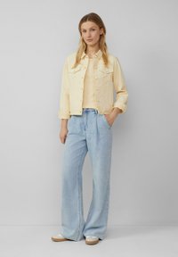  Lichtgele denimjack met twee borstzakken, gecombineerd met een beige top en hoge lichte blauwe wijde jeans. Casual schoeisel inbegrepen.