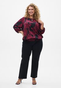 Blouse noire avec un motif floral rouge, manches froncées et décolleté en goutte d'eau, associée à un pantalon noir à jambes droites et des escarpins noirs à lanières.