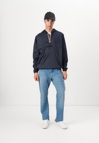 Marineblå vindjakke med zip og frontlomme, elastiske mansjetter og stripete detalj ved halsen, kombinert med lyseblå jeans og hvite joggesko.