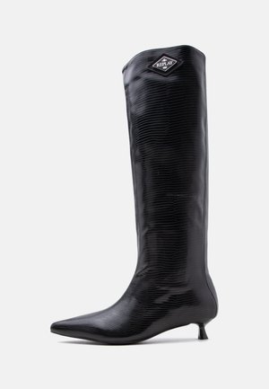 Botte noire montant jusqu’au genou avec un bout pointu, un petit talon kitten bas, une surface texturée et un écusson logo "REPLAY" près du haut.
