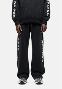 Zwarte katoenen hoodie met een voorzak en witte grafische tekst op de mouwen, gecombineerd met bijpassende zwarte joggingbroek in een losse pasvorm met zijprints.