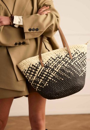 Persoon in beige blazer die een grote gevlochten boodschappentas vasthoudt met een zwart-natuurlijk patroon en bruine leren handvatten.