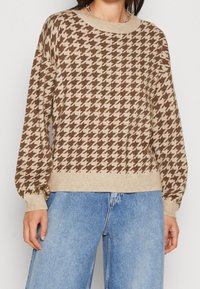 Brun och beige hounds-tooth-mönstrad stickad tröja med rund halsringning och ballongärmar, tillsammans med blå jeans.