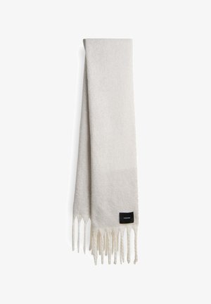 Zachte, lichte crèmekleurige sjaal van textuurstof, met franjes aan de randen en een klein zwart label met witte tekst.