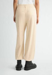 Beige weit geschnittene Hose mit elastischem Bund, die ein dezentes Allover-Logo-Muster und eine glatte Textur aufweist. Schwarze Knöchelstiefel werden getragen.