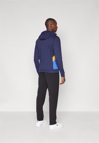 Marineblå hettegenser med et blått og gult geometrisk design på baksiden, sammen med svarte bukser og hvite sneakers på en lys bakgrunn.