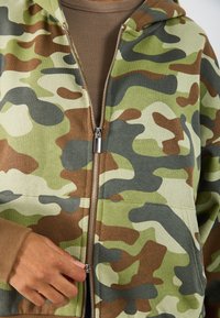 Sudadera con cremallera de camuflaje en verde, marrón y beige, con un tejido suave, bolsillos tipo canguro y herrajes plateados en la cremallera.