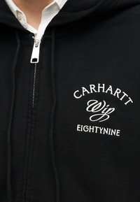 Musta vetoketjullinen huppari, jossa valkoinen kirjailtu teksti "CARHARTT Wip EIGHTYNINE" rinnassa, päällä valkoinen kauluspaita.