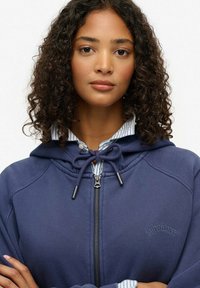 Sudadera azul marino con cremallera y cordón, que presenta un logotipo en el pecho y un cuello de camisa a rayas asomándose por debajo. Telas texturizadas visibles.
