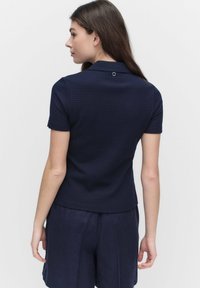 Meripaita tummansinisellä värillä, jossa on teksturoitu kuvio, lyhyet hihat ja pieni nappi selässä, yhdistettynä löysiin, tummiin shortseihin.