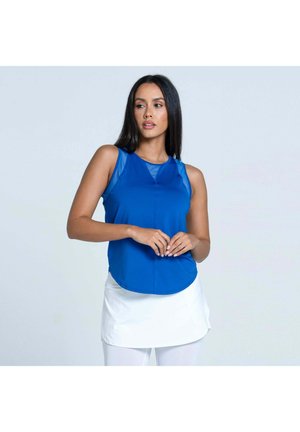 Frau mit langen dunklen Haaren, die ein ärmelloses blaues Oberteil und einen weißen Rock trägt, steht mit locker verschränkten Händen und schaut zur Seite.