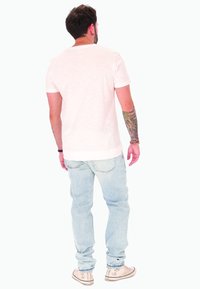 Uomo con un tatuaggio floreale sul braccio sinistro, indossa una maglietta bianca, jeans azzurri chiari e sneakers bianche, in piedi di spalle su uno sfondo bianco.