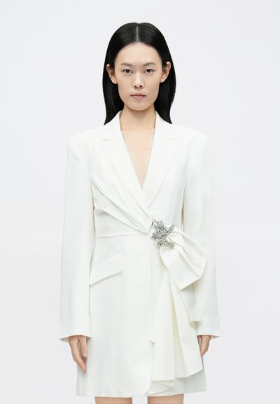 Robe blazer blanche à manches longues, col en V profond, poches latérales, et détail drapé à l'avant orné d'une broche argentée. Texture lisse.