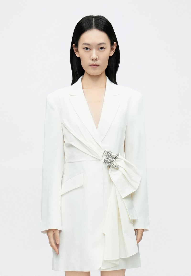 Vestido estilo blazer branco com mangas compridas, decote em V profundo, bolsos laterais e detalhe frontal drapeado realçado por um broche prateado. Textura suave.