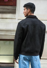Jachetă bomber neagră din suede fals, cu guler pufos, manșete și tiv ribbed, purtată cu blugi de culoare albastră deschisă. Vedere din spate.
