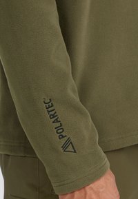 Pullover in pile verde oliva con texture liscia. Presenta il logo nero "Polartec" sulla manica. Design semplice con una forma aderente.