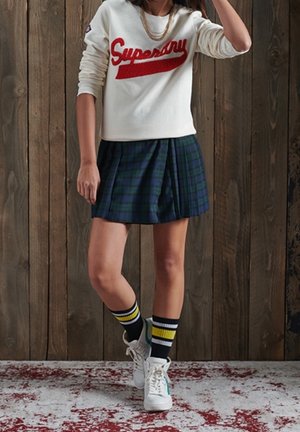 Sweat-shirt blanc avec le logo "Superdry" rouge, jupe plissée écossaise verte et marine, chaussettes montantes rayées et baskets blanches avec accent bleu.