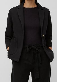 Blazer negro con bolsillos, cierre de un solo botón, corte entallado y tejido texturizado, combinado con una parte superior de canalé oscura y pantalones con cordón.