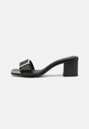 Black heeled mules with a glossy patent finish, square buckle detail, open toe design, and a medium block heel for stability.