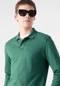 Cămașă polo verde cu mânecă lungă, guler și patch cu logo brodat. Modelul poartă ochelari de soare negri, cu o formă unghiulară, mărimea mare.