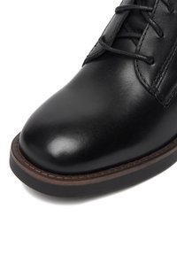 Černá kožená bota s hladkým povrchem, kulatou špičkou a kontrastní hnědou podrážkou. Obsahuje šněrování a prošívané detaily na svršku.