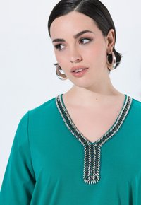 Top verde acqua con scollatura a V e dettagli intricati in perline lungo il collo, maniche corte e un tessuto liscio e texturizzato.