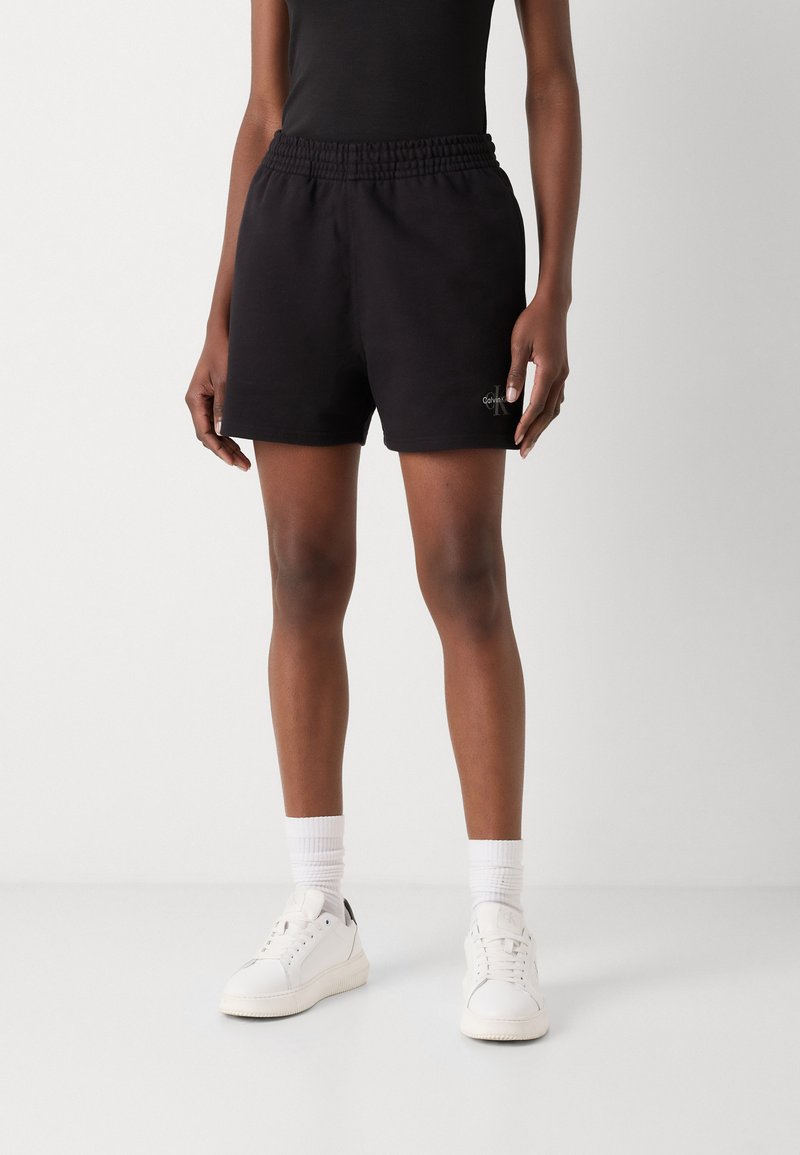 Zwarte katoenen shorts met een elastische tailleband, voorzien van een klein logo detail, gecombineerd met witte sneakers en geribbelde witte sokken.