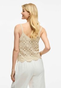 Top de encaje beige sin mangas con patrones de recortes circulares, dobladillo de puntilla y tirantes finos ajustables, combinado con pantalones blancos holgados.